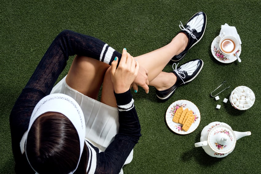 Trevor Paulhus' photograph for The Office of Angela Scott. The photo, taken from above, features a woman sitting on artificial grass wearing a pleated white tennis skirt, a blank and white preppy sweater, and a white visor. She also wears an interesting pair of black and white leather lace-up shoes. In front of her is a cup of tea on a saucer with a spoon and napkin, a plate of biscuits, a bowl of sugar cubes with a small set of tongs, and a teapot.